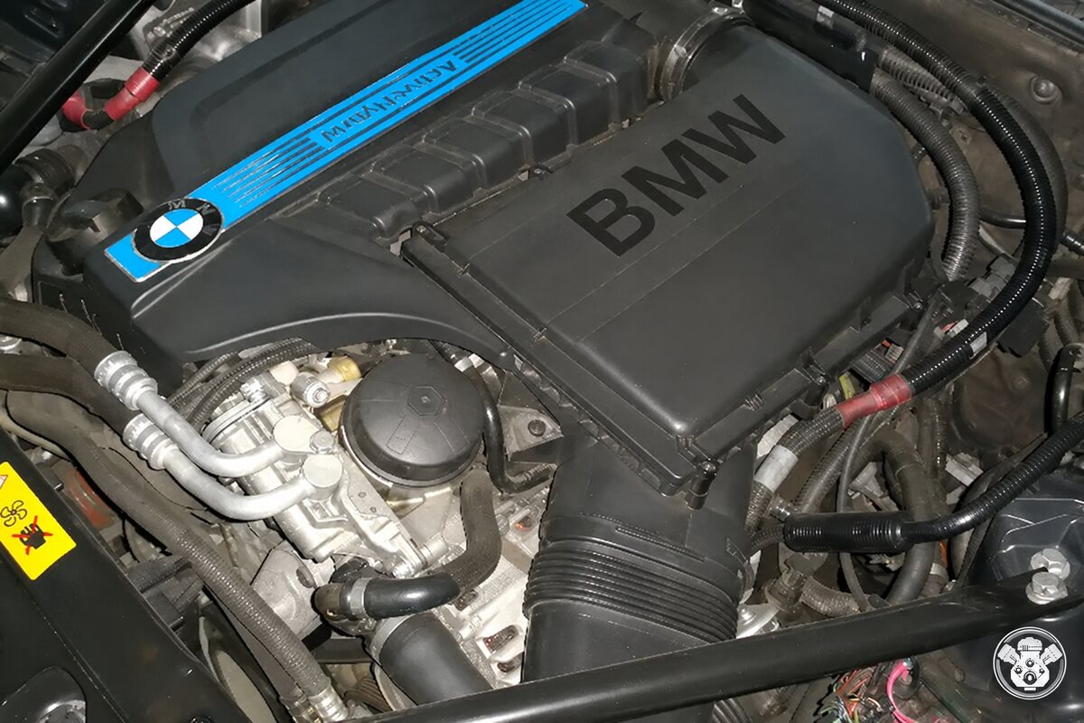 حجرة محرك BMW بغطاء أسود وشريط أزرق وشعار المصنع وأنابيب وأسلاك ظاهرة
